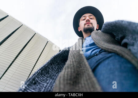 Ritratto di un elegante bel giovane con un rivestimento esterno. Basso angolo di visione di un uomo che indossa un cappotto e una sciarpa guardando fiduciosi. Foto Stock