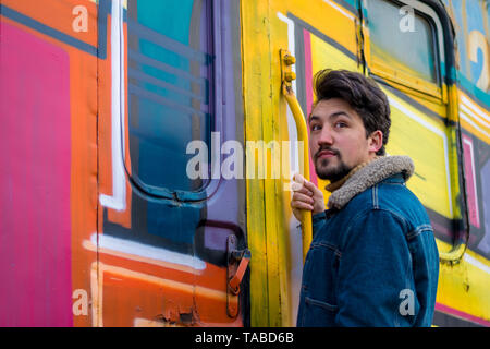 Ritratto di un elegante bel giovane con un denim giacca di lana all'aperto su di un treno. Un uomo che indossa un denim giacca di lana e un cappello, guardando la ca Foto Stock