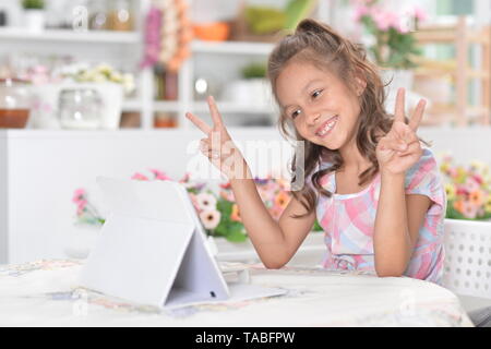 Ritratto di incantevole piccola ragazza seduta al tavolo della cucina nella parte anteriore del computer portatile e mostra segni di pace Foto Stock