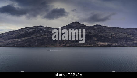 Una barca solitario che si muove attraverso l'acqua su un nuvoloso giorno contro lo sfondo di rotolamento di una gamma di montagna. Foto Stock