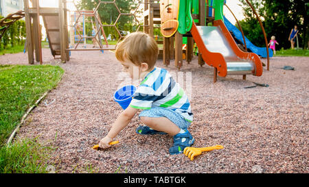 Ritratto di piccolo 3 anni bambino ragazzo seduto sul parco giochi e la sabbia di scavo con il giocattolo a forcella in plastica Foto Stock