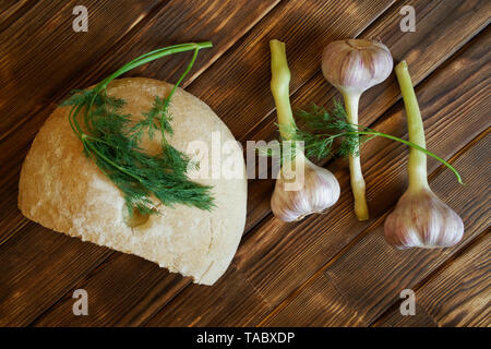 Aglio e rametti di aneto, insieme con una metà del bianco pane rotondo, giacciono su una tavola in legno rustico realizzato in tavole di pino. La luce diurna. Foto Stock