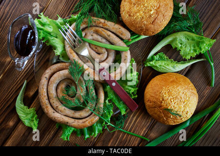 Salsicce alla griglia, mais panini, sauceboat trasparente e la forcella giacciono su una superficie in legno con verdi: insalata, aneto, cipolla e prezzemolo. Picnic - barbecue. Foto Stock