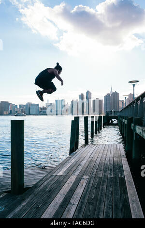 Stati Uniti d'America, New York, Brooklyn, il giovane uomo che fa parkour saltare da un palo di legno nella parte anteriore dello skyline di Manhattan Foto Stock