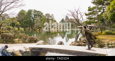 Una donna anziana che attraversa un laghetto su un ponte in il Kenroku-en giardino, Kanazawa, Giappone Foto Stock