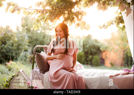 Donna incinta indossando elegante abito rosa in posa all'aperto con il tramonto sullo sfondo. La maternità. La maternità. Foto Stock