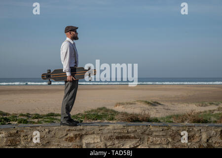 Uomo con longboard in piedi su una parete di fronte spiaggia e mare Foto Stock