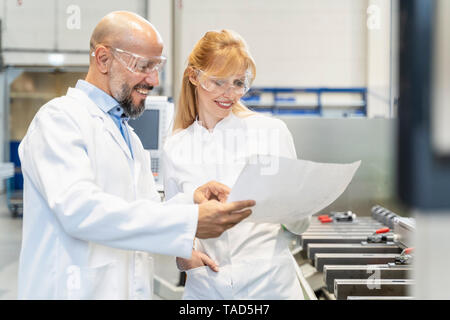 Due tecnici di felice indossando camici da laboratorio e occhiali di sicurezza guardando il piano in fabbrica Foto Stock