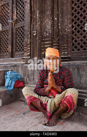 Donna anziana seduta con palme insieme in namaste saluto a Pashupati Mandir in Durbar Square, Bhaktapur, Valle di Kathmandu, Nepal Foto Stock