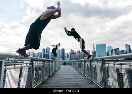 Stati Uniti d'America, New York, Brooklyn, tre giovani uomini a fare parkour salta sul molo di fronte skyline di Manhattan Foto Stock