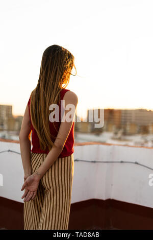 Vista posteriore della ragazza adolescente permanente sulla terrazza sul tetto della città al tramonto Foto Stock