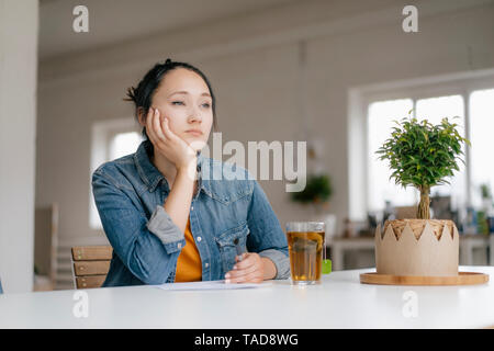Considerato giovane donna seduta a tavola con impianti e bicchiere di tè Foto Stock