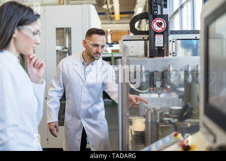 I colleghi indossando camici da laboratorio e occhiali di sicurezza guardando la macchina nel moderno stabilimento Foto Stock