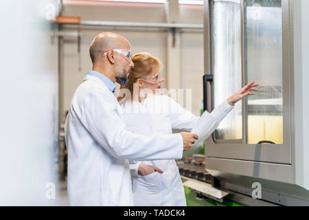 Due tecnici indossano camici da laboratorio e occhiali di sicurezza esaminando la macchina Foto Stock