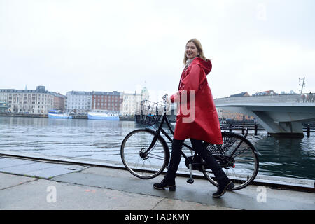 Danimarca, Copenaghen, donna felice spingendo biciclette presso il porto cittadino in condizioni di tempo piovoso Foto Stock