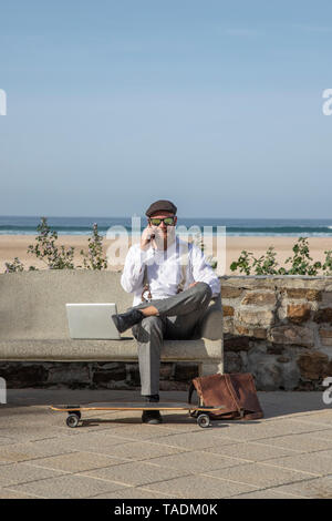 Uomo sorridente sulla seduta del telefono con il computer portatile su un banco di fronte spiaggia e mare Foto Stock