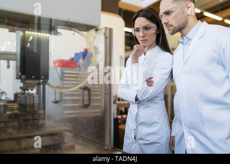 I colleghi indossando camici da laboratorio e occhiali di sicurezza guardando la macchina nel moderno stabilimento Foto Stock