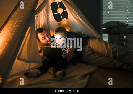 Padre e figlio condividendo una compressa in un buio tenda a casa Foto Stock