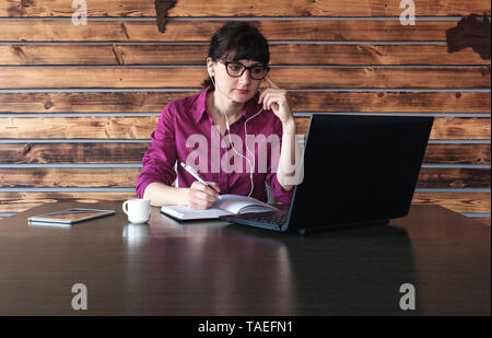 Imprenditrice ascoltando musica o un dettato come lavora presso il suo computer portatile prendere appunti in un moderno in legno ufficio accessoriato con co in primo piano Foto Stock