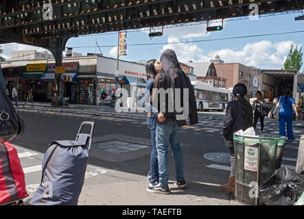 Una strada trafficata scena sotto la metropolitana sopraelevata con un paio di baciare, pedoni, bagagli e cestino. In Jackson Heights, Queens, a New York City. Foto Stock