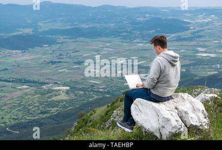 Giovane uomo seduto in cima alla montagna con vista di campi e colline e di lavoro on-line. Rete cellulare a banda larga il concetto di copertura. 5G. Foto Stock