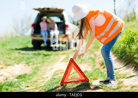 Giovane ragazza in arancione vest impostare il triangolo di breakdown sorge vicino a Broken auto, di strada Foto Stock