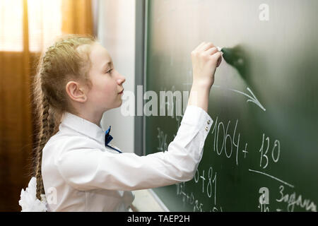 Il Little Schoolgirl detiene un gesso in mano e scrive una soluzione al problema della scuola di Blackboard Foto Stock