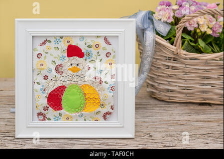 Un grande pollo con le uova di pasqua è ricamato e applicato. È situato in una cornice bianca. Merry decorazione per Pasqua nella parte anteriore di primule in ba Foto Stock