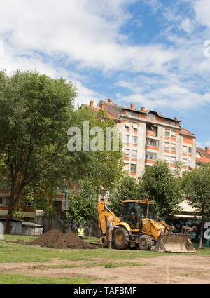 Escavatore lo scavo del terreno e uomo al lavoro nel parco tra gli edifici Foto Stock