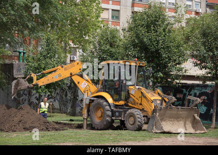 Escavatore lo scavo del terreno e uomo al lavoro nel parco tra gli edifici Foto Stock