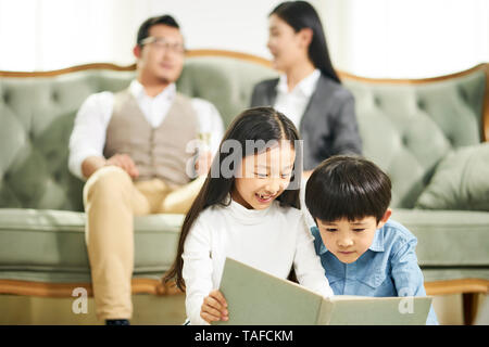 Due ragazzi asiatici del fratello e sorella seduta sul tappeto la lettura libro insieme a vivere in famiglia in camera con i genitori seduti sul divano in background. Foto Stock