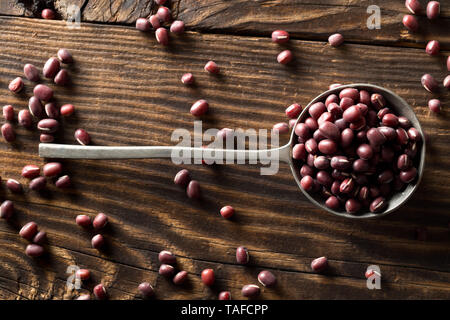 Materie, non cotte, essiccato adzuki (rosso) Mung bean in cucchiaio di metallo su tavola in legno rustico sfondo vista superiore piatta da laici al di sopra Foto Stock