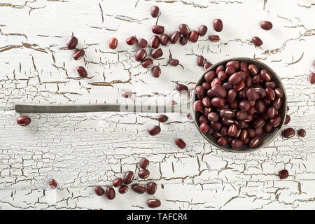 Materie, non cotte, essiccato adzuki (rosso) Mung bean in cucchiaio di metallo bianco su tavola in legno rustico sfondo vista superiore piatta da laici al di sopra Foto Stock