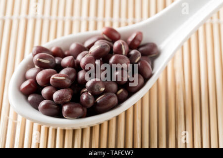 Materie, non cotte, essiccato adzuki (rosso) Mung bean in bianco cucchiaio sul tappetino di bambù sfondo con il fuoco selettivo Foto Stock