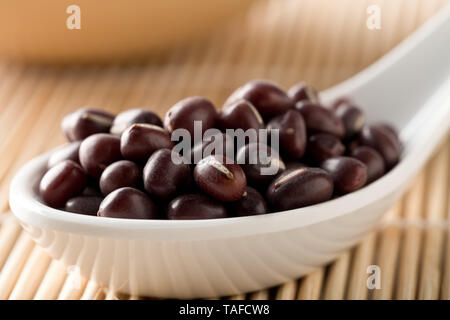 Materie, non cotte, essiccato adzuki (rosso) Mung bean in bianco cucchiaio sul tappetino di bambù sfondo con il fuoco selettivo Foto Stock