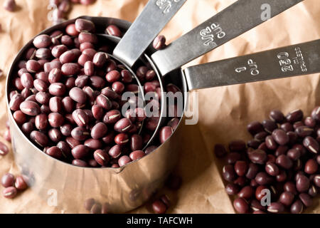 Materie, non cotte, essiccato adzuki (rosso) Mung bean in metallo tazze di misurazione su marrone carta da imballo sfondo con il fuoco selettivo Foto Stock