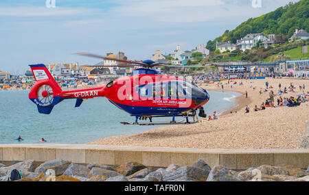 Lyme Regis, Dorset, Regno Unito. 25 maggio 2019. Devon Air Ambulance elicottero atterra sulla spiaggia affollata a Lyme Regis. L'elicottero decolla dall'affollata spiaggia con un paziente a bordo. Credito: Celia McMahon/Alamy Live News. Foto Stock