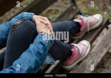 Donna seduta su una panchina, che tiene la gamba con le braccia, indossa jeans strappati neri e sneakers in tela rosa Foto Stock