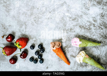 I frutti rossi come fragole ciliegie e mirtilli accanto a tre di gelati in cono su pietra chiara sfondo con copia spazio per il testo. Foto Stock