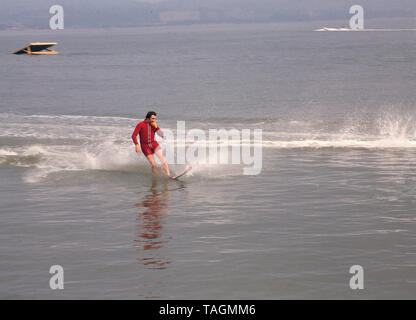 Anni Settanta, storico waterskier maschio in piedi sull'acqua a cavallo di un unico sci indossando una muta rosso avendo appena completato la sua corsa. Foto Stock