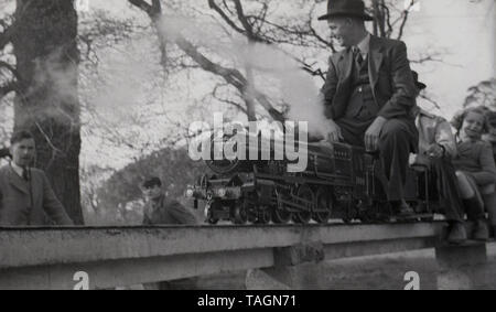 Degli anni Cinquanta, storico adatta uomo che indossa un cappello uomo che cavalca una miniatura del treno a vapore tirando i passeggeri su un evalated, sopra di terra via in un modello ferroviario, Inghilterra, Regno Unito. In questa epoca modello o delle ferrovie in miniatura con locomotive a vapore erano una popolare attrazione nei parchi e nelle località di mare e gestito da dedicati e gli appassionati di esperti. Foto Stock