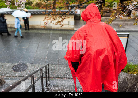 Uomo in rosso poncho passeggiando per le fasi durante il giorno di pioggia sulla strada marciapiede vicino Gion e caduti di fiori di ciliegio petali di fiori Foto Stock