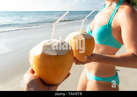 Close-up del giovane azienda noce di cocco con cannuccia in piedi presso la spiaggia vicino al mare Foto Stock