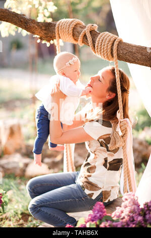 Donna sorridente holding baby ragazza seduta su altalena in legno all'esterno. Foto Stock