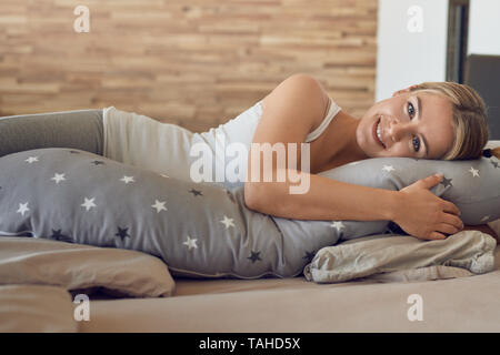 Felice sano giovane donna incinta in appoggio su un letto utilizzando uno speciale cuscino lungo come un supporto guardando la telecamera con un bel sorriso amichevole Foto Stock