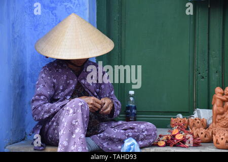 Il vietnamita vecchia donna con cappello conico vendere il suo negozio di souvenir sulle strade di Hoi An old town, Vietnam Foto Stock