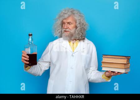 Vecchio con i capelli lunghi uomo che indossa Doctor's vestaglia studio isolato sulla parete blu in piedi che guarda alla bottiglia di whiskey in una sola mano azienda pila di libri in anot Foto Stock