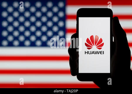 A profilarsi la mano di un uomo possiede uno smartphone visualizzando il logo della società cinese Huawei, con una bandiera degli Stati Uniti in background (solo uso editoriale). Foto Stock