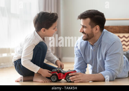 Felice padre e figlio giacente sul piano divertirsi giocando con il giocattolo auto racing insieme sorridente papà e bambino rilassarsi a casa impegnato nel gioco, ridere godeteveli Foto Stock