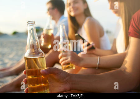 Gruppo di amici divertendosi gustando una bevanda rinfrescante e rilassante sulla spiaggia al tramonto. Giovani uomini e donne bere birra seduti su una spiaggia di sabbia in Foto Stock